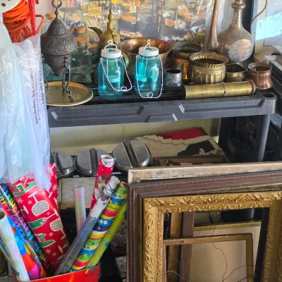 Sale Photo Thumbnail #304: Assortment of home goods, including vintage picture frames, rolls of wrapping paper, blue glass lanterns, and brass decor on plastic shelving.