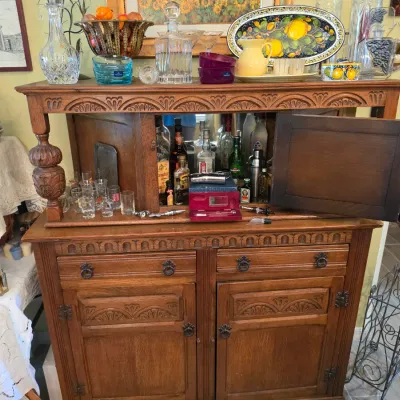 Sale Photo Thumbnail #71: Vintage carved oak dry bar cabinet with a drop-down mirror-backed mixing station, stocked with glassware and decanters.