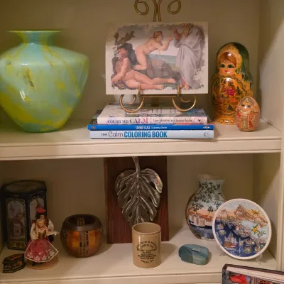 Sale Photo Thumbnail #227: Display shelves featuring decorative collectibles, including a swirl art glass vase, a set of Russian nesting dolls, adult coloring books, and souvenir ceramics.