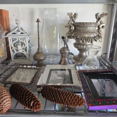Sale Photo Thumbnail #298: Collection of home decor items including decorative picture frames, glass vases, a cherub-adorned urn, brass vessels, and large pinecones.
