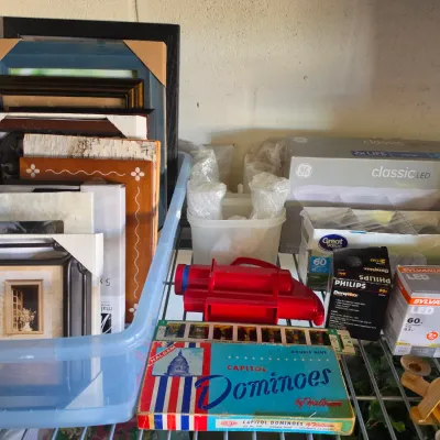 Sale Photo Thumbnail #300: Collection of assorted household goods including picture frames, lightbulbs, a vintage Halsam Capitol Dominoes set, and a red slide viewer on a wire shelf.