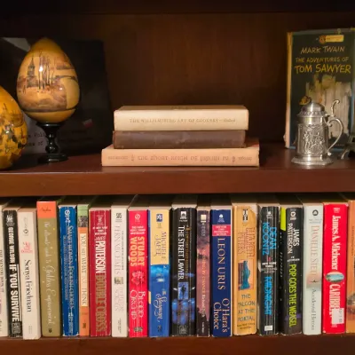 Sale Photo Thumbnail #217: Wooden bookcase filled with hardback and paperback books, featuring decorative items including painted egg ornaments, a miniature pewter tankard, and an Eiffel Tower figurine.