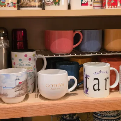 Sale Photo Thumbnail #23: Collection of souvenir and solid-color coffee mugs, travel tumblers, and a Godiva ceramic cup arranged on a cabinet shelf shelf-riser.