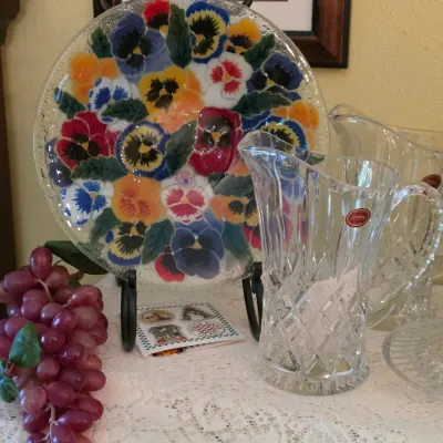 Sale Photo Thumbnail #79: Hand-painted glass plate featuring a colorful pansy design, accompanied by cut crystal glass pitchers and decorative home accents.