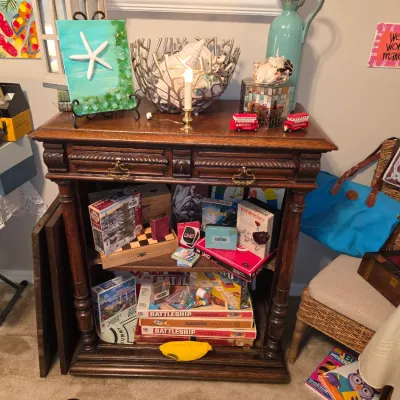 Sale Photo Thumbnail #171: Ornate carved wooden console table featuring lower open shelves filled with various vintage board games and puzzles, decorated on top with coastal-themed home decor.