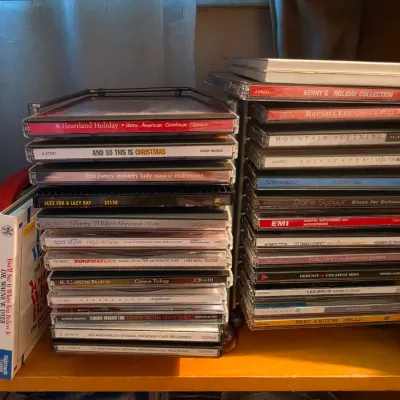 Sale Photo Thumbnail #116: Collection of music CDs in jewel cases stacked in wire storage racks on a wooden shelf, including holiday, jazz, classical, and pop titles.