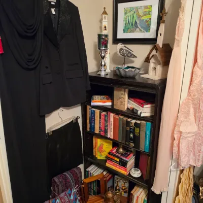 Sale Photo Thumbnail #191: Collection of women's vintage and modern clothing hanging beside a black wooden bookcase filled with books and decorative accents.