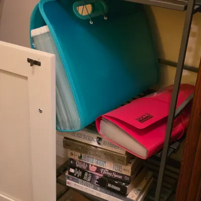 Sale Photo Thumbnail #226: Teal and pink plastic expanding file organizers resting on a small metal shelf above a stack of paperback and hardcover books.