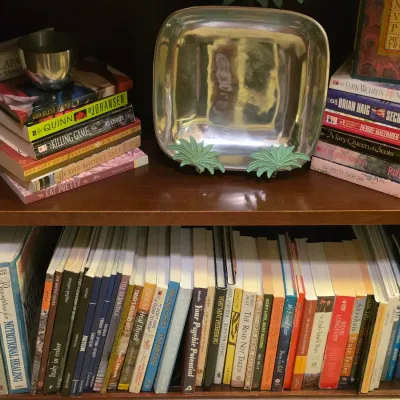 Sale Photo Thumbnail #249: Two wooden bookcase shelves filled with assorted fiction and non-fiction hardcovers and paperbacks, alongside a silver-tone tray display.