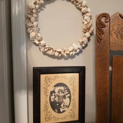 Sale Photo Thumbnail #189: Decorative seashell wreath hanging above a framed vintage-style portrait, set beside an ornate carved wood mirror frame.