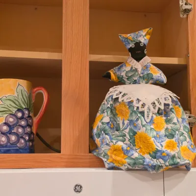 Sale Photo Thumbnail #31: Hand-painted ceramic grape-patterned pitcher alongside a handmade floral cloth toaster cover doll inside a wooden cabinet.
