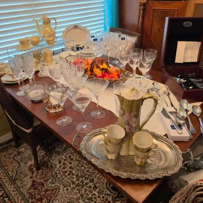 Sale Photo Thumbnail #101: Dining table display of fine glassware, crystal stemware, porcelain tea service, flatware set in a wooden chest, and a vintage hand-painted ceramic pitcher set on a silver-plated tray.