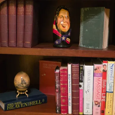 Sale Photo Thumbnail #216: Wooden bookcase filled with vintage hardcovers, paperbacks, a novelty caricature Russian nesting doll, and a decorative egg display.