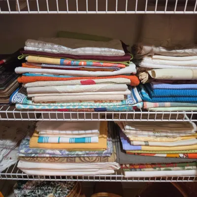 Sale Photo Thumbnail #67: Assorted vintage and modern home linens, including folded tablecloths, towels, lace pieces, and placemats arranged on wire closet shelving.