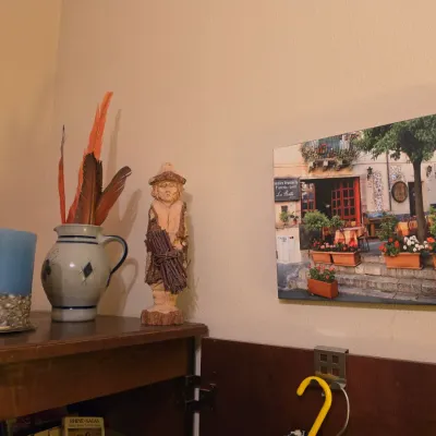 Sale Photo Thumbnail #251: Collection of decorative tabletop items, including a carved wooden figurine, a glazed ceramic pitcher holding feathers, a shell-embedded blue candle, and a canvas print of a European street scene.