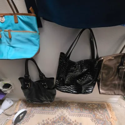 Sale Photo Thumbnail #165: Collection of women's handbags including a turquoise Michael Kors nylon tote, a black faux-croc tote, and a metallic bronze tote hanging on a wall.