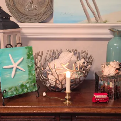 Sale Photo Thumbnail #173: Collection of coastal-themed decor items including a sea glass and starfish canvas artwork, silver coral bowl filled with shells, blue ceramic pitcher, and small red London bus collectibles on a wooden tabletop.