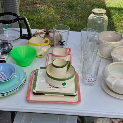 Sale Photo Thumbnail #329: Assortment of vintage housewares including colorful ceramic dishes, glassware, planters, and a glass coffee carafe arranged on an outdoor table.