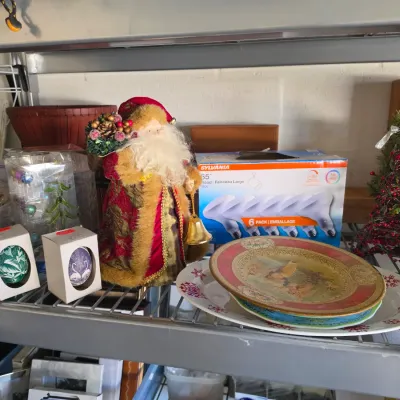 Sale Photo Thumbnail #299: Collection of holiday decorations, featuring a plush Father Christmas figure, decorative plates, boxed ornaments, and a mini beaded Christmas tree arranged on metal shelving.