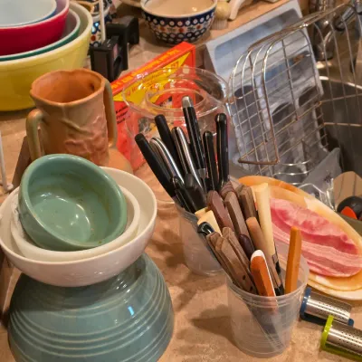 Sale Photo Thumbnail #13: Collection of vintage kitchenware including stacked Pyrex and ceramic mixing bowls, a pottery pitcher, and plastic cups holding various cutlery and utensils.