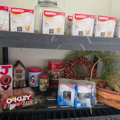 Sale Photo Thumbnail #282: Black plastic storage shelves containing light bulbs in boxes, holiday tins, a small ceramic house, an Easter basket with decorative bunny, and boxed Oakley E Frame goggles.