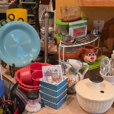 Sale Photo Thumbnail #15: Collection of colorful kitchen items including a blue Fiestaware-style plate, ceramic pitcher, novelty face mug, salad spinner, and small baking dishes.
