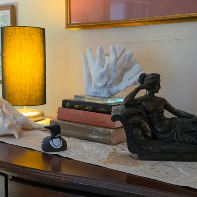 Sale Photo Thumbnail #138: Collection of tabletop decor including a classical dark figure statue reclining on a couch, white coral specimen on acrylic base, conch shells, and a small table lamp with a dark shade.
