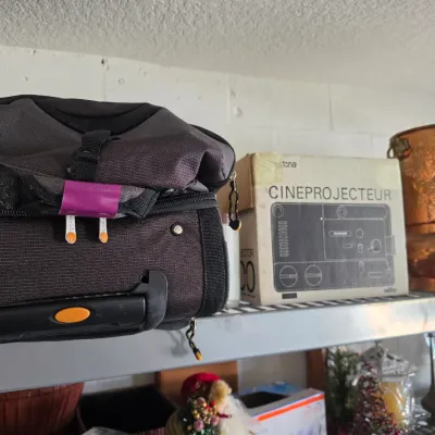 Sale Photo Thumbnail #296: Selection of shelf items featuring a black rolling travel bag, a vintage Keystone Cineprojecteur box, and a hammered copper urn.
