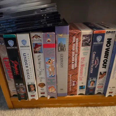 Sale Photo Thumbnail #117: Collection of vintage VHS tapes featuring classic movies and special interest titles alongside a stack of CD jewel cases on a wooden shelf.