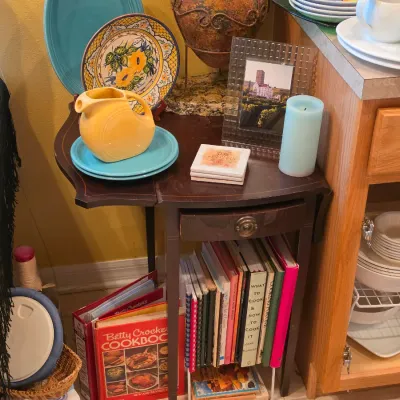 Sale Photo Thumbnail #40: Vintage drop-leaf side table displayed with a yellow Fiesta pitcher, decorative ceramic plates, a collection of vintage cookbooks, and glassware.
