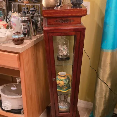 Sale Photo Thumbnail #12: Tall, narrow wooden curio display cabinet with glass door panels, housing decorative collectibles alongside kitchenware and teacups displayed on an adjacent counter.