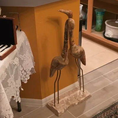 Sale Photo Thumbnail #9: Pair of carved stone or composite bird sculptures mounted on metal legs and a rectangular stone base, alongside a lace-covered table displayed with ceramic dishware.
