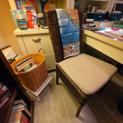 Sale Photo Thumbnail #244: Dark woven rattan side chair with a beige upholstered seat cushion, draped with a printed map scarf, shown alongside a woven basket filled with books.