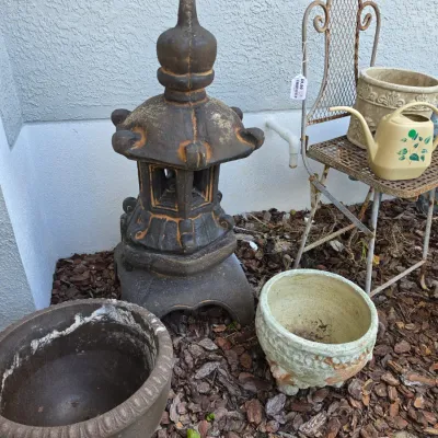 Sale Photo Thumbnail #349: Collection of garden decor including a concrete pagoda lantern statue, wrought iron plant stand, ceramic planters, and a small watering can.