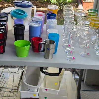 Sale Photo Thumbnail #322: Assortment of modern kitchenware and glassware displayed on folding tables, including plastic pitchers, travel mugs, margarita glasses, and organization bins.
