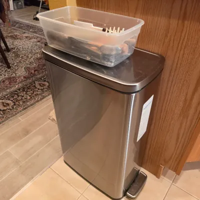 Sale Photo Thumbnail #21: Stainless steel step-on trash can topped with a clear plastic storage container holding kitchen utensils.