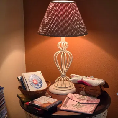 Sale Photo Thumbnail #112: White open-work wire table lamp with an open-weave dark fabric shade, displayed on a round side table with books, CDs, and small decorative baskets.