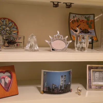Sale Photo Thumbnail #239: Collection of decorative tabletop objects displayed on white shelves, including picture frames, crystal decor, an acrylic scorpion paperweight, and ceramic plates.