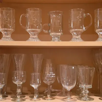 Sale Photo Thumbnail #24: Collection of assorted vintage glassware arranged on two wooden cabinet shelves, including etched Irish coffee mugs, champagne flutes, cordial glasses, and wine goblets.