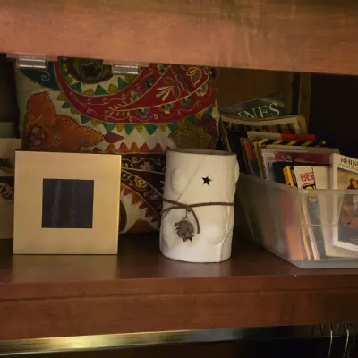 Sale Photo Thumbnail #254: Wooden shelf stored with mixed decorative items, including a colorful paisley embroidered pillow, small framed pictures, a white ceramic star luminary candle holder, and a clear plastic bin filled with books.