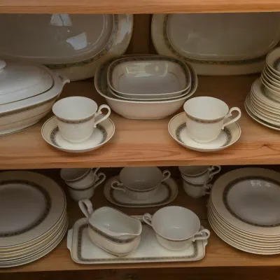 Sale Photo Thumbnail #99: Extensive fine china dinnerware service featuring a delicate floral border and gold rims, displayed on wooden shelves.
