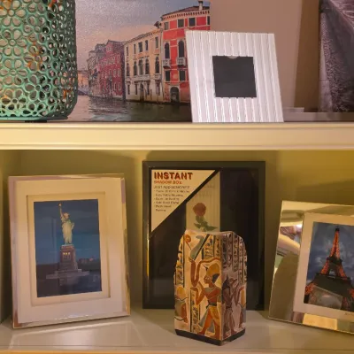 Sale Photo Thumbnail #235: Collection of framed artwork and home decor items, including travel photographs of the Statue of Liberty and Eiffel Tower, a Venice canvas print, and an Egyptian-themed vase displayed on white shelving.