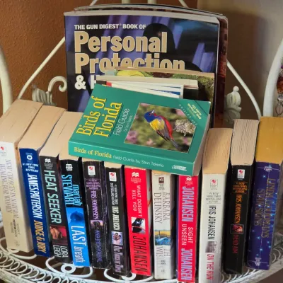 Sale Photo Thumbnail #144: Collection of assorted paperback and hardcover books arranged on a white wire rack, featuring authors such as Iris Johansen and James Patterson alongside field guides.
