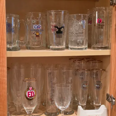 Sale Photo Thumbnail #19: Collection of assorted beer glasses and steins, featuring brands like Beck's, Warsteiner, Hacker-Pschorr, and Berliner Kindl, displayed on cabinet shelves.