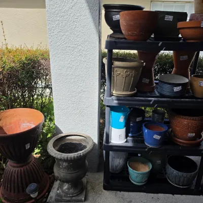 Sale Photo Thumbnail #344: Collection of assorted ceramic, terracotta, and concrete garden planters arranged outdoors on black plastic shelving units and on the ground.