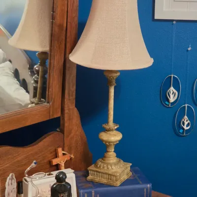 Sale Photo Thumbnail #188: Traditional distressed cream table lamp with a fabric bell shade, resting on a wooden dresser alongside framed perfume bottles and decor.