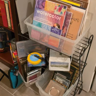 Sale Photo Thumbnail #258: Black metal wire rolling rack holding clear plastic bins filled with art supplies, instruction books, and picture frames.