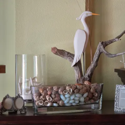 Sale Photo Thumbnail #85: Coastal-themed table décor featuring a carved white egret on driftwood, glass vessels filled with seashells and glass stones, picture frames, and a wall crucifix.