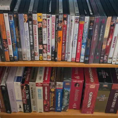 Sale Photo Thumbnail #124: Two wooden bookshelf shelves filled with a collection of DVDs and VHS tapes, featuring popular film and fitness titles.