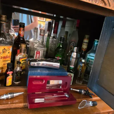 Sale Photo Thumbnail #204: Selection of barware and liquor bottles displayed inside a carved wooden hutch, featuring a stainless steel cocktail shaker, bottle stoppers, and various spirits.